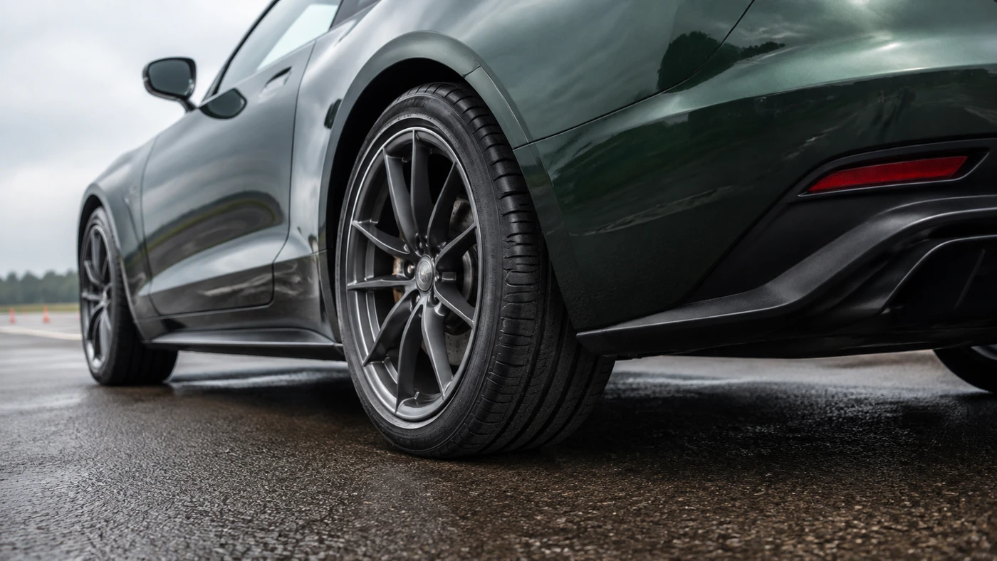 Rear wheel and treaded tire of a dark green coupe contacting damp asphalt on a test pad