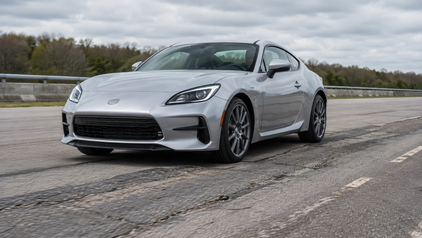 Stiffer Suspension explainer showing a silver sports coupe on patched asphalt at a closed test facility