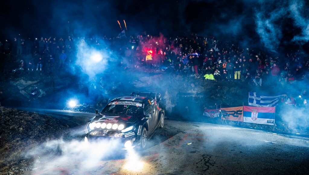 WRC rally car driving at night through a snowy stage with spectators